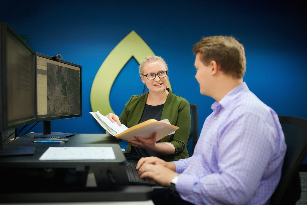 A standing woman shows a folder to a man seated at a workstation