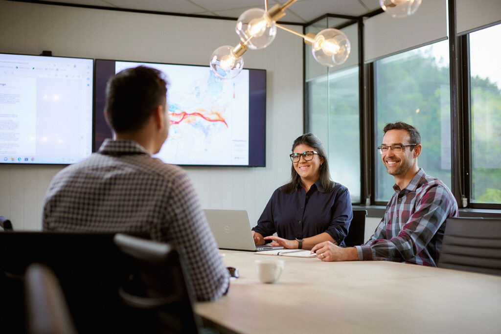 Three people around a conference table with a laptop and a large display on the wall