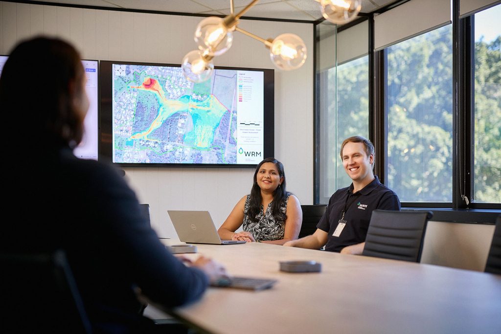 Three people around a conference table with a laptop and a large display on the wall